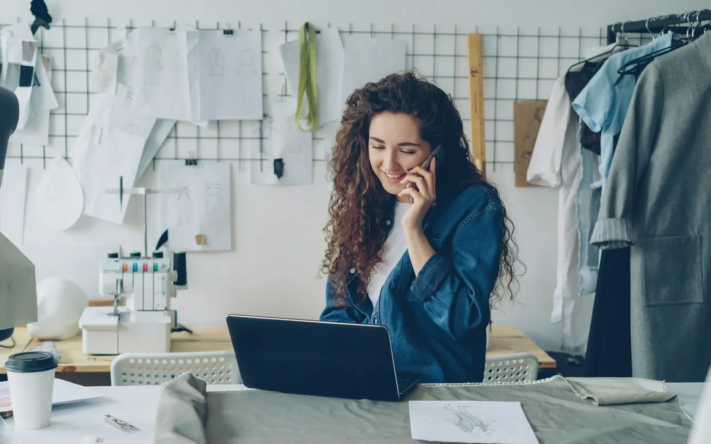A shop owner smiling on the phone while working on a laptop in a tailoring studio