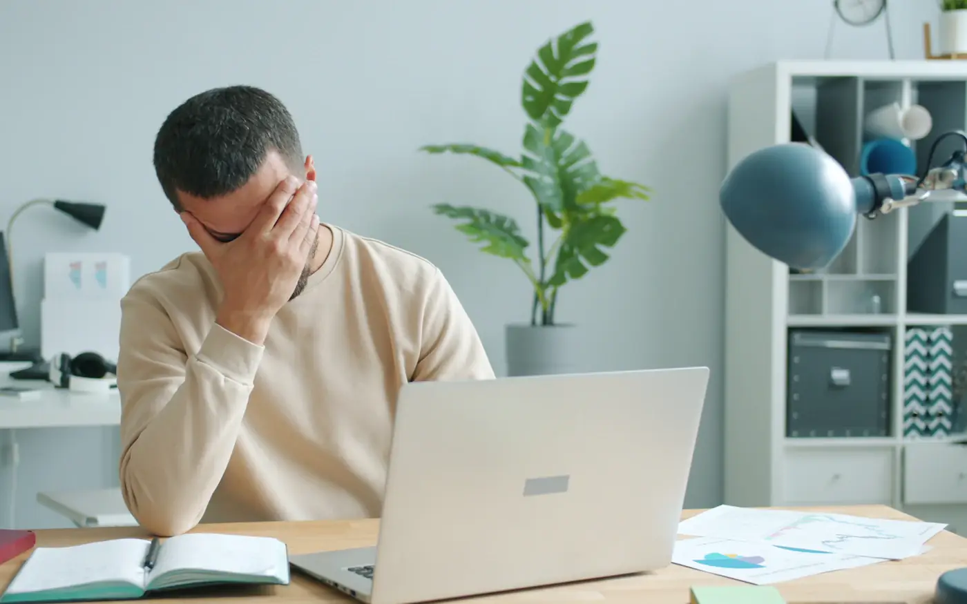 A tired man rubbing his face in front of a laptop at a desk